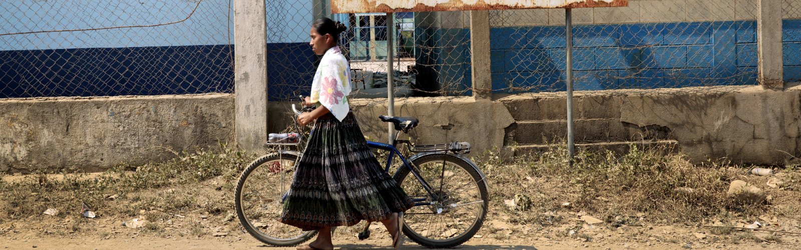 A Q'eqchi' woman with a bicycle walks down the unpaved main road through Sepur Zarco.
